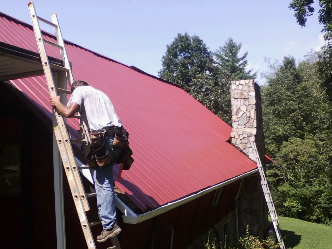 Roofer on a ladder beside a red metal roof and stone chimney — Soffit & Fascia Repair by Roofing Brothers in Chisago City